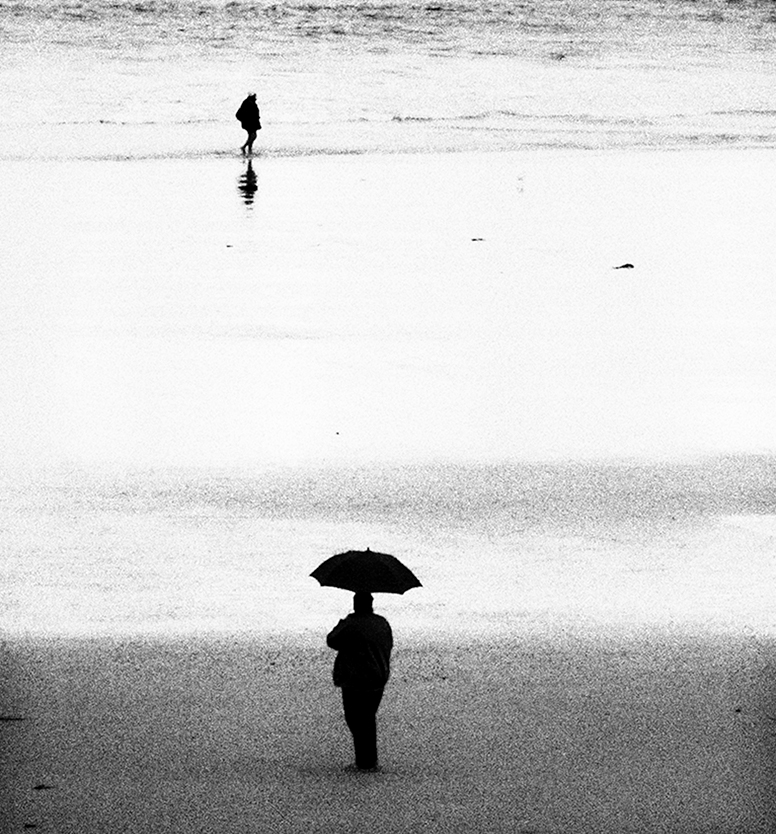 Photo noir et blanc d'un homme de dos tenant un parapluie sur une plage. Dans le fond, une femme marche dans l'eau